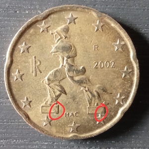 May include: A close-up of a gold-colored 2002 Euro coin. The coin features a stylized image of a person and stars around the edge. The coin has the letter "R" and the text "2002" on it.
