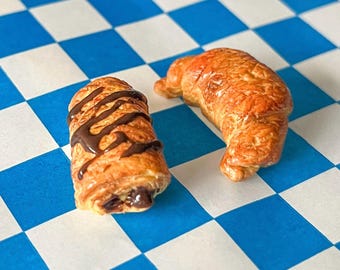 May include: Two miniature pastries on a blue and white checkered surface. One is a croissant, golden brown with a curved shape. The other is a chocolate pastry, with chocolate drizzle. The background is a blue and white checkered pattern.