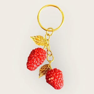 May include: A gold-coloured keyring featuring two red raspberry charms with gold-coloured leaves. The keyring has a gold ring and chain, creating a charming and fruity accessory.