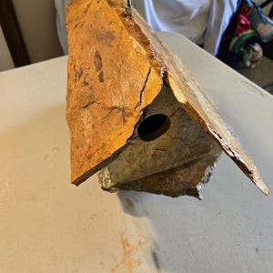 May include: A rustic birdhouse made from natural stone, with a warm, earthy colour scheme of browns and tans. The birdhouse has a circular entry hole and a metal hook for hanging. The roof is angled, providing shelter.