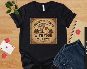 May include: Black t-shirt with a poker-themed graphic. The design features playing cards, poker chips, and the text "I BOUGHT THIS SHIRT WITH YOUR MONEY!!" in a vintage style. The shirt is displayed on a wooden surface.