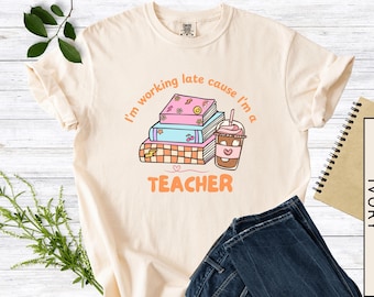 Könnte beinhalten: Elfenbeinfarbenes T-Shirt mit einer Grafik von gestapelten Büchern, einem Kaffeegetränk und dem Text "I'm working late cause I'm a teacher". Das Design ist in Pastellfarben gehalten. Eine Jeans ist sichtbar.