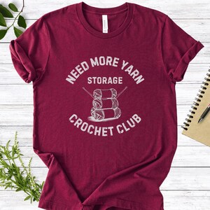 Puede incluir: Camiseta granate con el texto blanco "Need More Yarn Storage Crochet Club". El gráfico presenta tres ovillos de lana con agujas de tejer. La camiseta está sobre una superficie de madera blanca con vegetación.