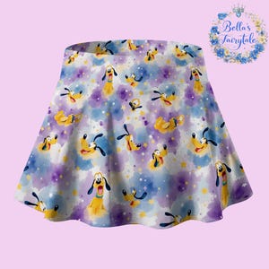 May include: A flared skirt featuring a repeating pattern of Pluto the dog on a watercolor background of purple, blue, and white. The skirt has a wide waistband and is suitable for various occasions. The text "Bella's Fairytale" is in a floral wreath.