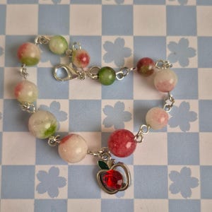 Op de afbeelding: Een armband met ronde kralen in rood, groen en wit. De armband is verbonden door een zilveren ketting en heeft een bedel in de vorm van een rode edelsteenappel.