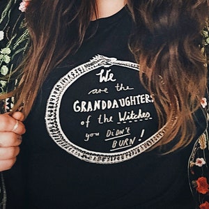 May include: A woman wearing a black t-shirt with a white circle graphic that says "We are the Granddaughters of the Witches you Didn't Burn!" The graphic is surrounded by a snake design. She is wearing a black jacket with floral embroidery.