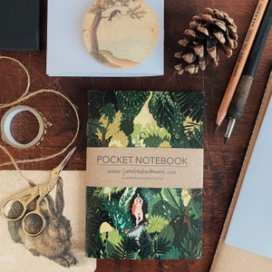 May include: A small green and brown pocket notebook with a colorful illustration of a woman standing in a forest. The notebook cover has the text "POCKET NOTEBOOK" and "www.sandradiechmann.com" printed on it. The text "ILLUSTRATED & MADE IN THE UK" is also printed on the cover.