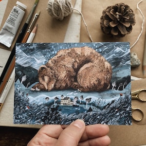 以下が含まれることがあります： 冬の風景の中で眠っている茶色のクマのイラストが描かれたポストカード。山、木、家がある風景の中で休んでいるクマが描かれています。ポストカードは手で持たれており、周囲には画材や自然の要素があります。