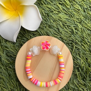 Puede incluir: Una pulsera colorida con cuentas planas y redondas en tonos rosa, amarillo y blanco. La pulsera está adornada con dijes de conchas marinas y una pequeña flor. Se muestra sobre un círculo de madera, con una flor blanca en el fondo.