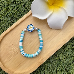 Op de afbeelding: Een armband met schijfvormige kralen in wit, lichtblauw en blauw. Het heeft een blauw-witte bloemvormige bedel, goudkleurige kralen en parels. De armband wordt gepresenteerd op een houten dienblad, naast een witte bloem.