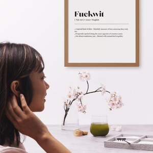 Op de afbeelding: Een houten lijst met het woord "Fuckwit" en de definitie ervan. Een vaas met kersenbloesems en een glas groene vloeistof staan op een tafel. Een persoon staat op de voorgrond.