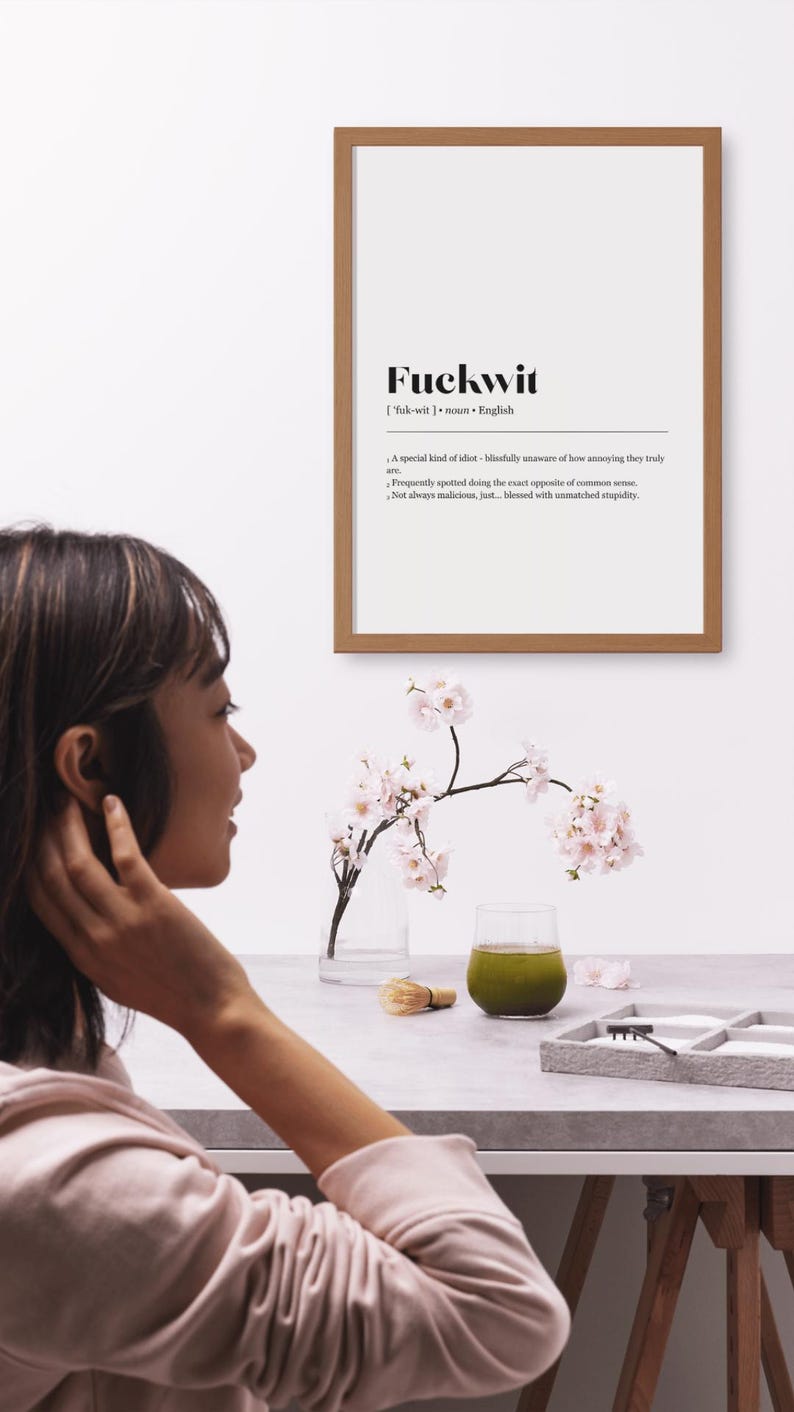 Op de afbeelding: Een houten lijst met het woord "Fuckwit" en de definitie ervan. Een vaas met kersenbloesems en een glas groene vloeistof staan op een tafel. Een persoon staat op de voorgrond.