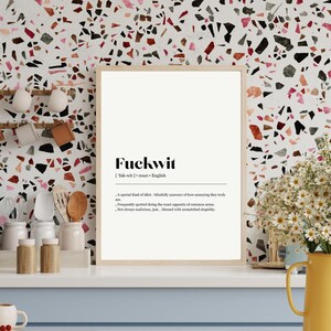 Op de afbeelding: Een ingelijste print met het woord "Fuckwit" en de definitie ervan in zwarte tekst. De print staat op een witte achtergrond en wordt weergegeven op een wit aanrecht. Rechts staat een gele kan met bloemen.