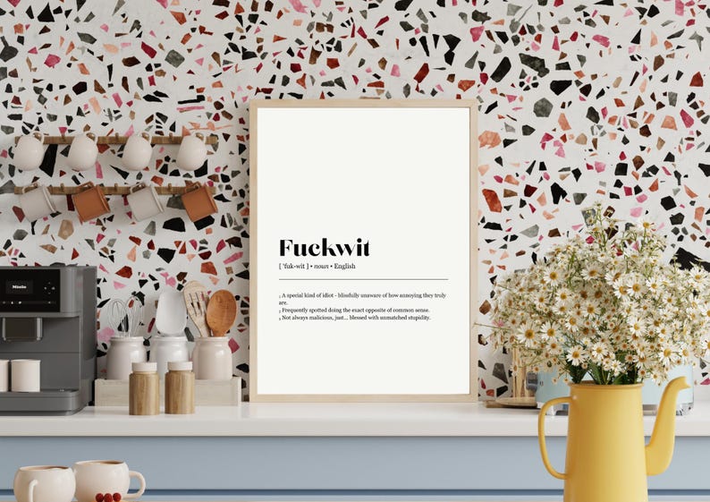 Op de afbeelding: Een ingelijste print met het woord "Fuckwit" en de definitie ervan in zwarte tekst. De print staat op een witte achtergrond en wordt weergegeven op een wit aanrecht. Rechts staat een gele kan met bloemen.