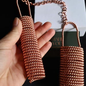 Puede incluir: Dos objetos cilíndricos de color cobre, hechos a mano, conectados por una cadena. Los objetos tienen un asa en forma de lazo y están hechos de alambre enrollado firmemente. Los objetos se sostienen en una mano sobre un fondo blanco y oscuro.