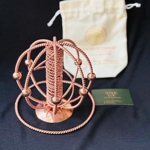 May include: A copper-colored metal sculpture with a spherical design, featuring interconnected rings and spheres. A small, beige drawstring bag and a green business card are in the background. The sculpture has a circular base and a central vertical element.