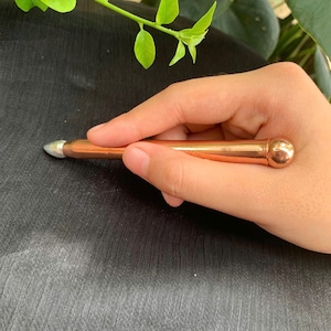 May include: A copper-colored massage tool with a rounded end and a pointed tip. The tool is held in a hand against a dark background, with green leaves in the upper left corner. The tool is approximately 5 inches long.