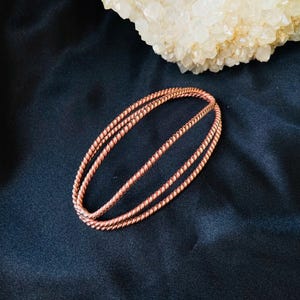May include: Two copper-colored, twisted-wire bracelets are displayed on a dark blue surface. The bracelets are oval-shaped and have a textured appearance. A large, light-colored crystal formation is in the background.