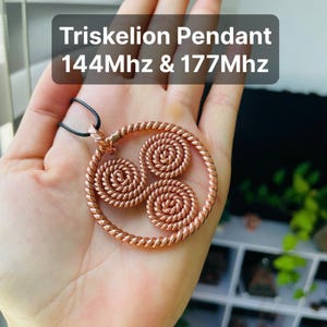 May include: Copper-toned Triskelion pendant featuring three spiral motifs within a circular frame, suspended from a black cord. The image includes text: "Triskelion Pendant 144Mhz & 177Mhz".