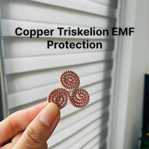 Puede incluir: Un triskel en cobre, símbolo de tres espirales, se sostiene sobre un fondo blanco. El texto "Copper Triskelion EMF Protection" se muestra encima del triskel. El triskel es de color marrón rojizo.