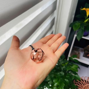 Puede incluir: Un colgante de esfera de cristal facetado de color rosa, suspendido de un cordón negro. El cristal se sostiene en una palma abierta, mostrando su intrincado diseño y color. El colgante es un accesorio decorativo.