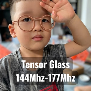 Peut inclure: Un enfant porte des lunettes rondes de couleur cuivre avec un design en corde torsadée. Les lunettes ont une esthétique vintage. Le texte "Tensor Glass 144Mhz-177Mhz" est affiché en blanc sur un fond sombre.