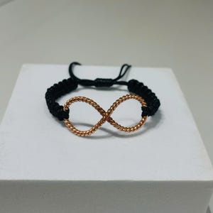 Op de afbeelding: Een zwart gevlochten armband met een koperkleurig oneindigheidssymbool. Het oneindigheidssymbool is gemaakt van gedraaid metaal. De armband wordt op een wit oppervlak gepresenteerd.