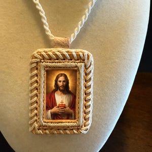 May include: A rectangular pendant featuring an image of Jesus Christ, framed with woven beige and white cord. The pendant hangs from a white braided cord. The image depicts Jesus with a red robe and a glowing heart.