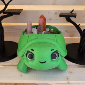 May include: A bright green 3D-printed turtle-shaped planter with large black eyes and a smiling mouth. The shell contains several upright crystal points in shades of orange, pink, and gray, nestled among small white stones inside the hollow cavity.