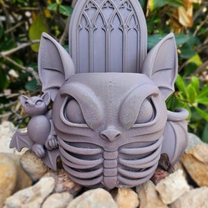 May include: A gray, 3D-printed planter shaped like a bat-faced creature with large eyes, bat wings, and a ribcage design. A smaller bat figure is on the left side. The planter has a gothic arch-shaped back and sits on a bed of rocks.