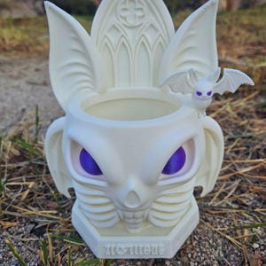 May include: A white, gothic-style planter with a bat-like design. The planter features purple eyes, bat wings, and a small bat figurine. The base has the word "TOTENE" in a stylized font. The planter is designed for decorative use.