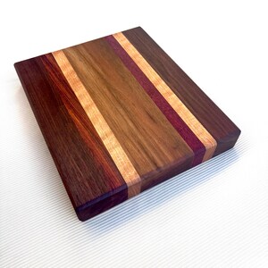 Op de afbeelding: Een rechthoekige houten snijplank met een gestreept ontwerp. De plank heeft afwisselende strepen van donkerbruin, lichtbruin, middenbruin en paars hout. De snijplank is gemaakt van hardhout en is geschikt voor gebruik in de keuken.