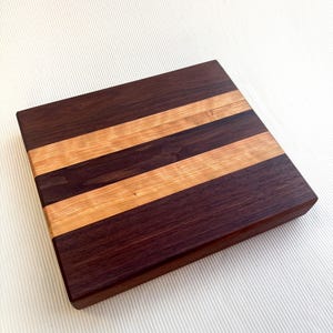 Op de afbeelding: Een rechthoekige houten snijplank met afwisselende donkerbruine en lichtbruine strepen. De houtnerf is zichtbaar en de plank heeft een glad, afgewerkt oppervlak. Dit keukenartikel is geschikt voor het bereiden van voedsel.