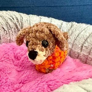 May include: A handmade brown and tan crocheted dog with black eyes and a black nose. The dog is wearing an orange collar and is resting on a pink fluffy pillow inside a grey and white plush dog bed.