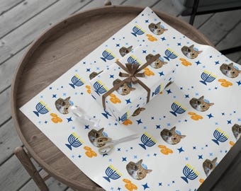 May include: White wrapping paper with a repeating pattern of cat faces wearing yarmulkes, blue menorahs, orange dreidels, and blue stars. A wrapped gift with a brown ribbon sits on the paper, along with a pair of scissors.