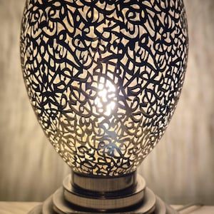 Op de afbeelding: Een eivormige tafellamp met een donkere, ingewikkelde metalen schaal met uitsparingen waardoor licht schijnt. De lamp staat op een ronde, getrapte metalen voet met een gladde, reflecterende afwerking die een warme gloed verspreidt.