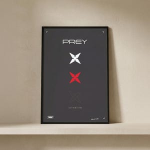 Könnte beinhalten: Schwarz gerahmtes Poster mit dem Wort "PREY" in Weiß. Darunter befinden sich drei stilisierte X-Formen, die mittlere in Rot. Der Text "HUNT OR BE HUNTED" steht unten. Das Poster ist auf einem weißen Regal ausgestellt.