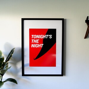 May include: A framed art print with the text "TONIGHT'S THE NIGHT" in white on a red background, with a black diagonal shape. A black baseball cap hangs on the wall to the right.