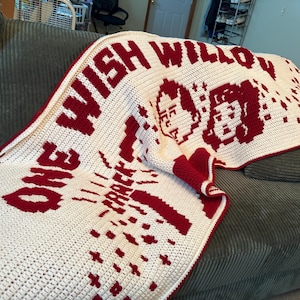 May include: A cream-colored crochet blanket featuring dark red pixelated designs. The text reads ONE WISH WILLOW and CRACK. The graphic includes two pixelated faces and scattered star-like shapes, all rendered in a dark red yarn on a cream background.