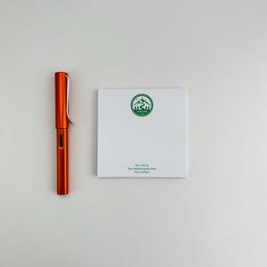 May include: An orange pen with a silver clip and a white notepad with a green mountain logo and the text "CUSTOM". The notepad also includes the text "Your Name", "Your address goes here", and "Your contact".