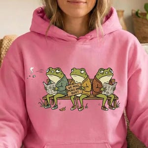 Könnte beinhalten: Ein hellrosa Kapuzenpullover mit einer Grafik von drei grünen Fröschen, die auf einer Holzbank sitzen. Die Frösche tragen Pullover und lesen Zeitungen, während der mittlere Frosch ein Schild mit der Aufschrift WE MISS PEACE AND QUIET hält.