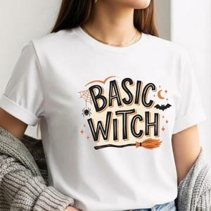 Könnte beinhalten: Ein weißes Kurzarm-T-Shirt mit dem schwarzen Schriftzug BASIC WITCH. Das Design enthält orangefarbene und schwarze grafische Elemente wie ein Spinnennetz, eine Spinne, eine Mondsichel, eine fliegende Fledermaus, kleine Sterne und einen Besen.