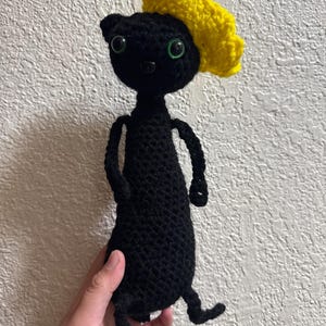 May include: A handmade crochet doll featuring a solid black body with a slender neck and torso. The doll has two bright green plastic eyes, a small black nose, and a textured, vibrant yellow crocheted beret-style hat attached to its head.