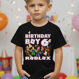 以下が含まれることがあります： 「BIRTHDAY BOY 6」の文字とRobloxのキャラクターがプリントされた黒いTシャツ。誕生日のお祝いにぴったりの、風船と紙吹雪を使ったカラフルなデザインです。男の子は青いジーンズを履いています。