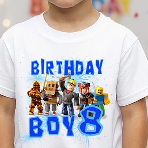 Puede incluir: Camiseta blanca con un gráfico azul que dice "BIRTHDAY BOY 8" y presenta personajes de dibujos animados. Los personajes son de varios colores y diseños, incluyendo robots y figuras con herramientas.