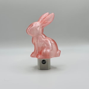 May include: A pink, iridescent bunny night light. The bunny is sitting upright with its ears perked up. The night light base is a neutral gray color.