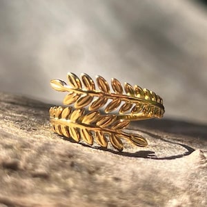 Peut inclure: Une bague dorée avec un motif de feuilles. La bague s'enroule autour du doigt avec deux bandes de feuilles détaillées. La bague est posée sur un fond en bois naturel, mettant en valeur le design et la couleur complexes du bijou.
