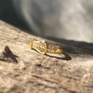 Peut inclure: Une bague dorée avec un motif de boucle, incrustée de petits pierres précieuses scintillantes. La bague est posée sur une surface en bois naturelle et texturée, avec des ombres.
