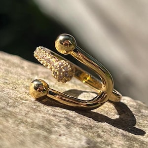 Op de afbeelding: Een goudkleurige ring met een uniek ontwerp. De ring heeft een gebogen band met twee bolvormige accenten en een gedeelte bezet met kleine, sprankelende stenen. De ring is geplaatst tegen een natuurlijke, getextureerde achtergrond.