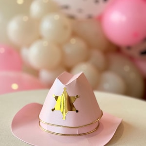 Op de afbeelding: Een lichtroze cowboyhoed met een gouden sheriffster en gouden rand. De hoed is gemaakt van papier, waarschijnlijk voor een feestje. Roze en witte ballonnen zijn op de achtergrond te zien.